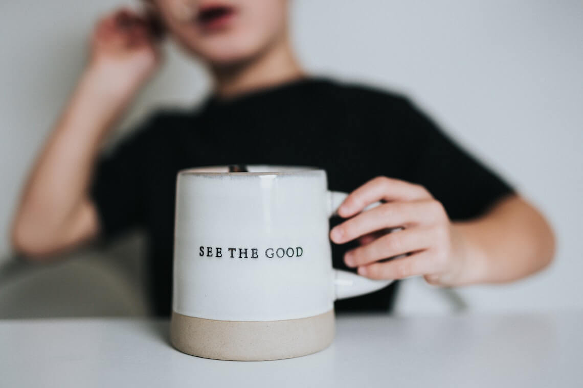 A mug that says see the good is being held by a child that is out of focus behind it