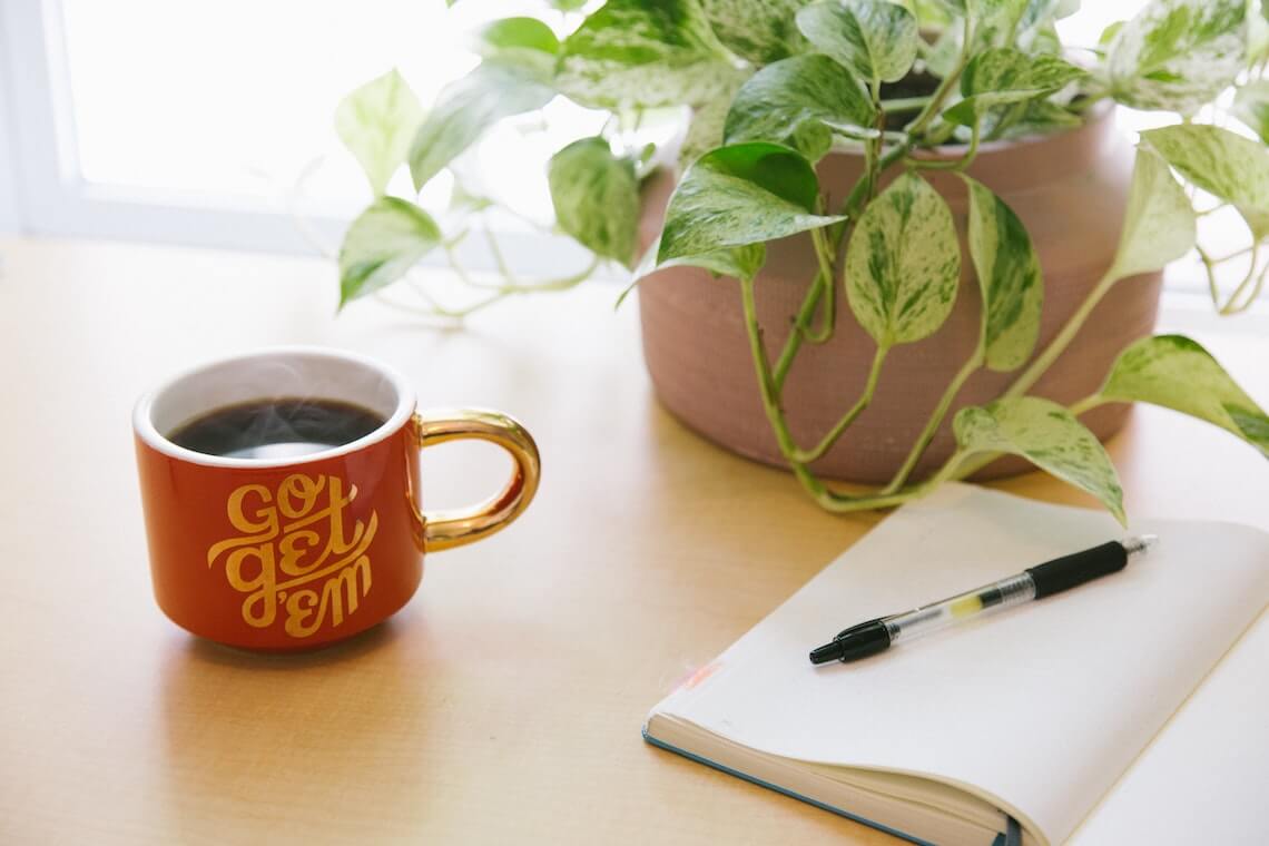 A cup of coffee in a mug that says go get em is set upon a wooden tabletop with an empty notebook next to it with a pen on top 