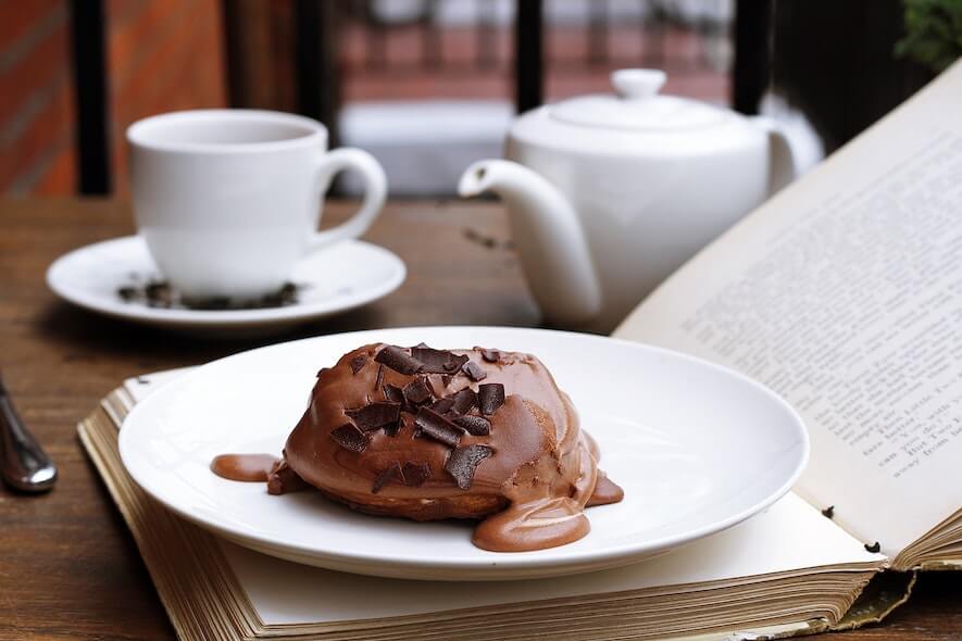 A chocolate dessert sits atop a white plate, with a cup, saucer, and teapot blurry in the background