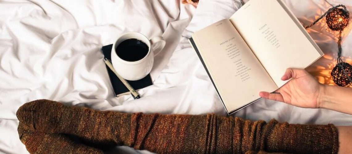 A person is reading a book in bed with a cup of coffee nearby, only the person's legs are visible